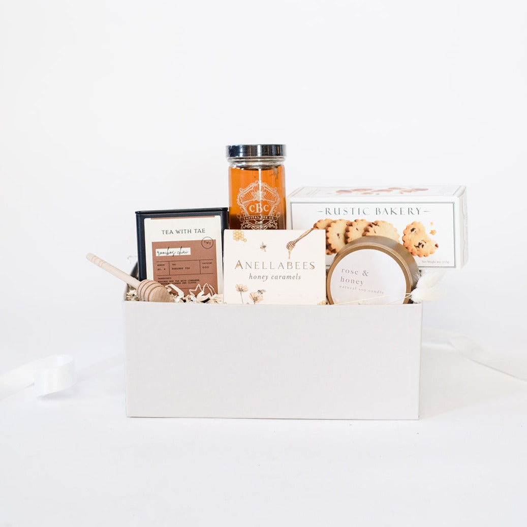 Light grey box containing wooden honey drizzler, black Rooibos chai tea tin, jar of raw capital bee co. raw holly honey, box of Anellabees honey caramels, a box of rustic bakery pecan shortbread cookies and a rose & honey candle in a gold tin.