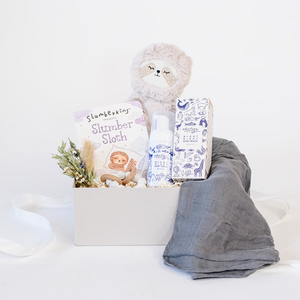 A light gray gifting box with a dried floral bundle, Slumberkins Slumber Sloth and hardback book, Chelsey Wang night night baby wash, a charcoal muslin swaddle and silicone and wooden teether.