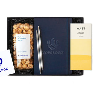 White custom logo card, vanilla popcorn with custom logo, custom logo navy notebook with strap, set of two silver pens, Large mast hazelnut chocolate bar in yellow and white packaging. All in black gifting box with black paper shred