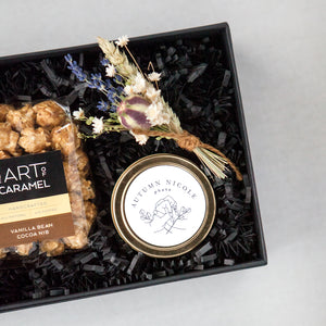 Small black gifting box with black paper shred, the art of caramel popcorn in clear packaging with black, tan, and brown sticker. Gold candle tin with client logo, floral bundle with lavender and white flowers