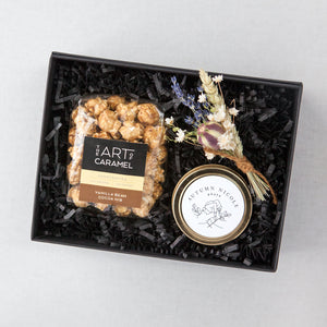 Small black gifting box with black paper shred, the art of caramel popcorn in clear packaging with black, tan, and brown sticker. Gold candle tin with client logo, floral bundle with lavender and white flowers