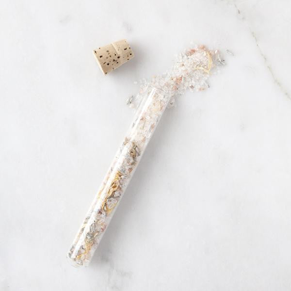 Cork sitting next to an open glass vial with bath salts and dried flowers spilling out onto a marble surface. 