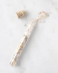 Cork sitting next to an open glass vial with bath salts and dried flowers spilling out onto a marble surface. 