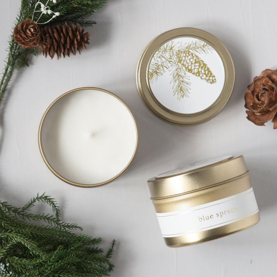 blue spruce natural hand-poured soy candle, hand-poured in a 4 oz. brushed gold metal tin. The image shows three of them: one open (with a white wax candle contained inside), one showing the candle's top part (a blue spruce branch in gold on a white label), and one showing the candle sideways (with a white label featuring gold lettering) surrounded by Christmas decor on a white background. 