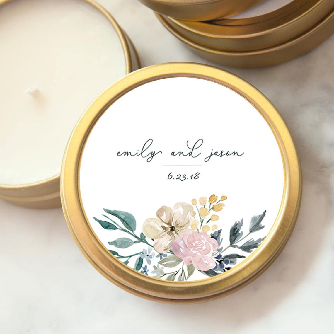  Our Custom wedding favor candle Emily, featured in tis gold tin. The names “emily and jason” are written in a flowing, handwritten-style script,  The date "6.23.18" is in clean font. The floral design consists of a blend of soft, pastel colors, including pink, cream, blue, and green tones, executed in a loose watercolor style
