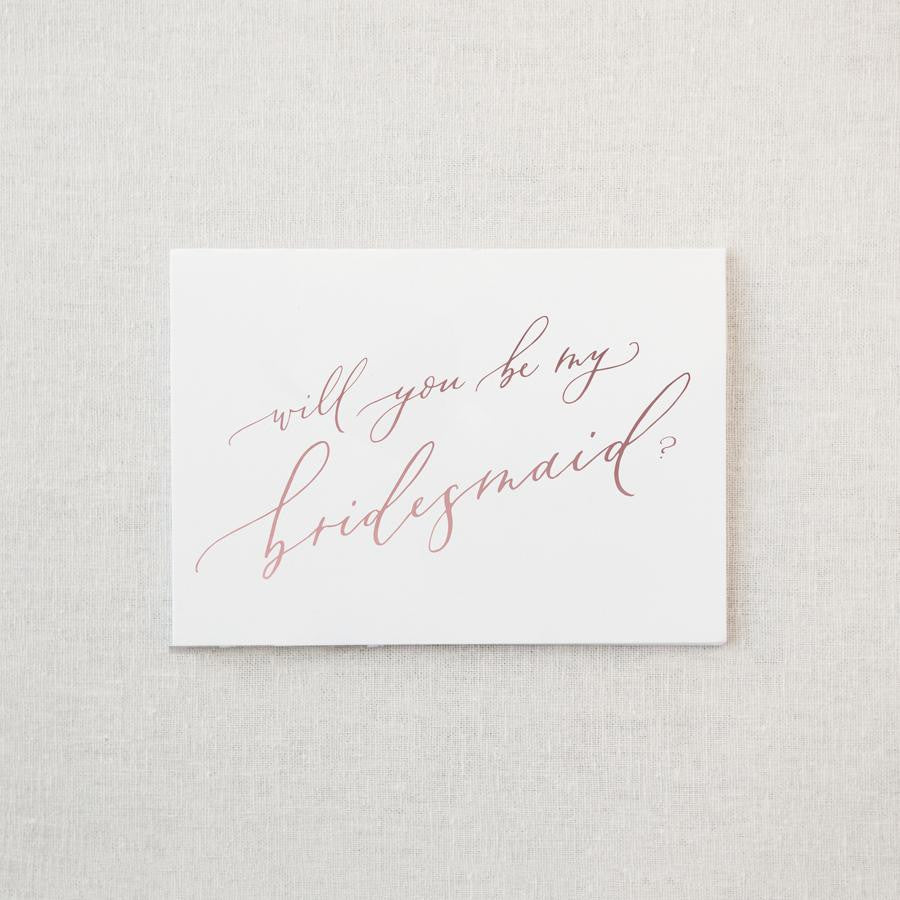 White envelope size card (approx. 3.5" x 5"), reads, "Will you be my bridesmaid?" in rose gold text.
