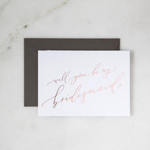 White envelope size card (approx. 3.5" x 5"), reads, "Will you be my bridesmaid?" in rose gold text. Slate gray envelope option behind. 