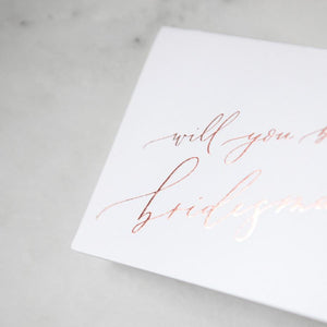 White envelope size card (approx. 3.5" x 5"), reads, "Will you be my bridesmaid?" in rose gold text zoomed in.