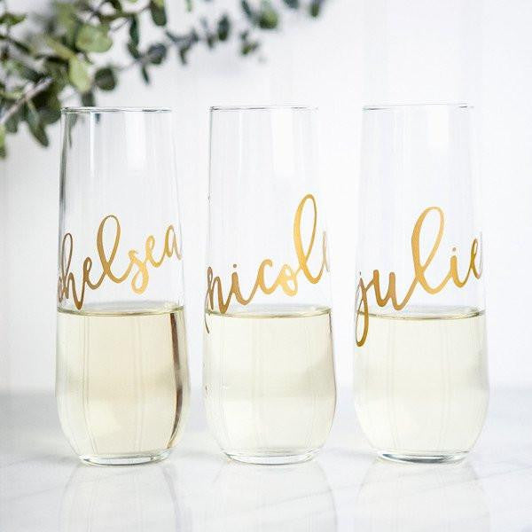 Three stemless flute glasses half half-filled with champagne gold script names (Chelsea, Nicole, and Julie). In a white background with a green branch on the background.