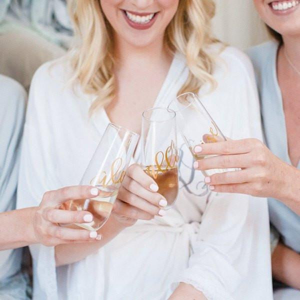 Image of a bride and two bridesmaids holding their champagne flutes personalized in gold lettering. 
