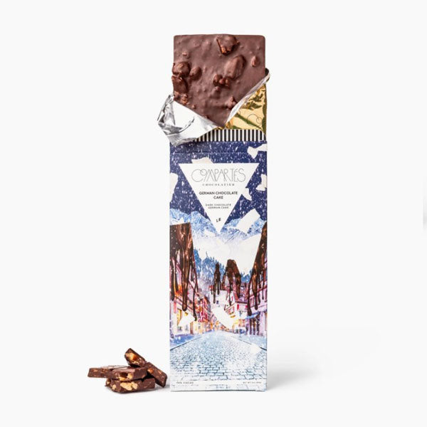 Compartes german chocolate cake bar featured in its wrapping that has the image of a Christmas village semi opened showing the inner gold wrapping and the brown chocolate bar and three pieces of the chocolate next to it. 