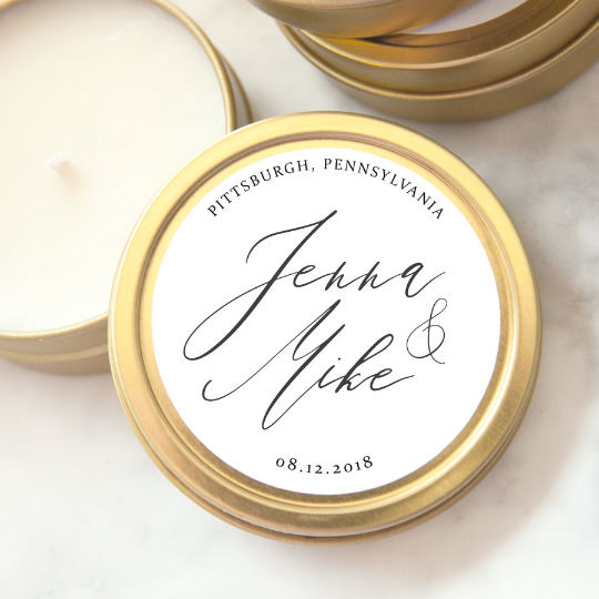 A close-up photograph displays a gold-colored candle tin featuring personalized details. a candle tin made of gold-colored metal, with a round shape and a visible seam. Inside the tin, there is a white candle. A circular label covers the top, displaying elegant calligraphy. The label features the names "Jenna &amp; Mike," the location "Pittsburgh, Pennsylvania," and the date "08.12.2018."