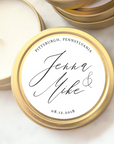 A close-up photograph displays a gold-colored candle tin featuring personalized details. a candle tin made of gold-colored metal, with a round shape and a visible seam. Inside the tin, there is a white candle. A circular label covers the top, displaying elegant calligraphy. The label features the names "Jenna & Mike," the location "Pittsburgh, Pennsylvania," and the date "08.12.2018."