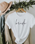 Custom script bride tee image shown hanged in a black clothing rack in front of a beige cardigan jean jacket and beige hat with black detail and green plant wrapped around. 