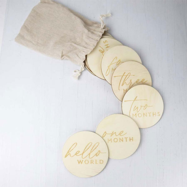 First year milestone discs  presents a set of engraved wooden milestone discs for a baby, along with a small drawstring bag, arranged on a neutral, textured surface. The discs are arranged in a slight arc, creating a visually appealing composition that guides the viewer's eye. The words “hello world”, “one month”, “two months”, “three”, “four” and "six months" are visible, along with others partially visible.