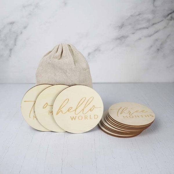 The image showcases a collection of wooden milestone discs for a baby, along with a small canvas bag, arranged on a light-colored surface,arranged on a solid, light-colored surface, likely a tabletop, with a light-colored marble or stone backdrop.