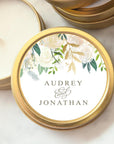 Close up image of Floral Candle custom wedding favor candle gold metal  container with a circular shape and a lid. On the lid, there is a white label that is decorated with floral arrangements.  The floral arrangement has white flowers and green leaves. Containers the name Audrey and Jonathan.  The candle is opened showing the white candle inside with another container on the upper right in a marble-like background. 