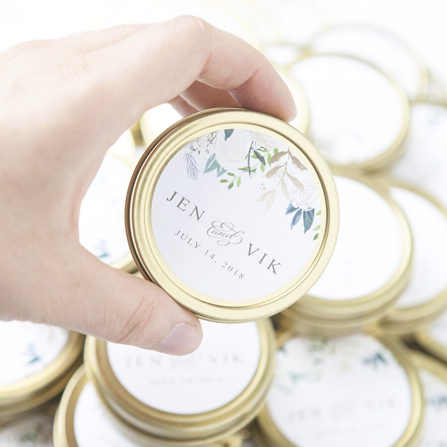 The image showcases a hand holding a gold-rimmed container with a personalized label for a wedding, surrounded by several similar containers,It's made of gold metal with a circular shape and a lid. On the lid, there is a white label that is decorated with floral arrangements. The label contains the names "Jen" and "Vik" in a stylish script font, beneath which the date "July 14, 2018" is printed. The floral arrangement has white flowers and green leaves. 