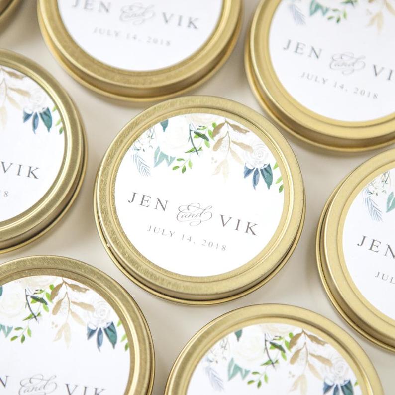 A close-up photograph presents several small, round, gold-colored containers, each adorned with a floral-themed label featuring the names "Jen &amp; Vik" and the date "July 14, 2018."