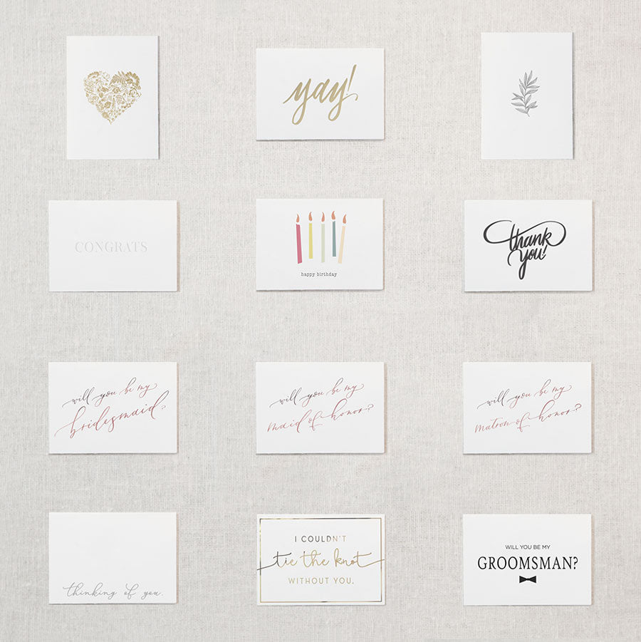 Display of twelve white cards. Gold floral heart, "Yay!" in gold foil, grey botanical branch, "congrats" embossed, "happy birthday" written under five multicolored candles, "thank you!" in grey script, "will you be my bridesmaid?" in rose gold, "will you be my maid of honor?" in rose gold, "will you be my matron of honor?" in rose gold, "thinking of you" in grey script, "I couldn't tie the knot without you" in gold foil, "will you be my groomsman?" in black text