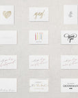 Display of twelve white cards. Gold floral heart, "Yay!" in gold foil, grey botanical branch, "congrats" embossed, "happy birthday" written under five multicolored candles, "thank you!" in grey script, "will you be my bridesmaid?" in rose gold, "will you be my maid of honor?" in rose gold, "will you be my matron of honor?" in rose gold, "thinking of you" in grey script, "I couldn't tie the knot without you" in gold foil, "will you be my groomsman?" in black text