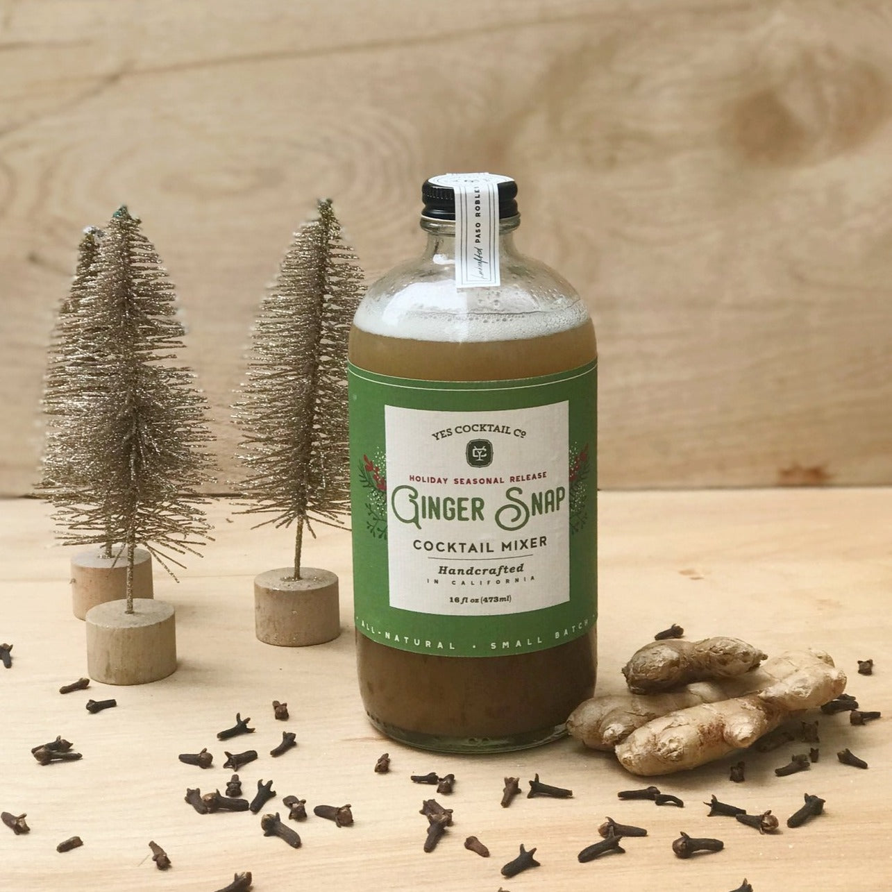 Clear glass bottle with black cap, white sticker seal and green label next to decorative snow dusted trees fresh ginger and cloves. Ginger Snap cocktail mixer.