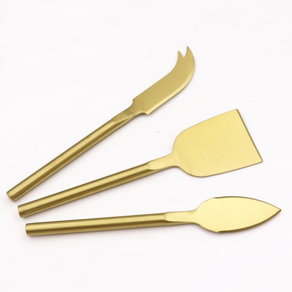 Matte gold cheese knives, set of three, gold-plated stainless steel, one pronged knife for hard cheeses, one wedge knife for semi-hard cheeses, and one tear-shaped knife for soft cheeses. Shown  in a white background.  