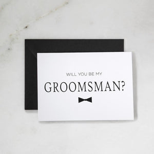 White envelope size card (approx. 3.5" x 5"), reads, "Will you be my groomsman?" in black text, with a bowtie graphic. Black envelope option pairing.
