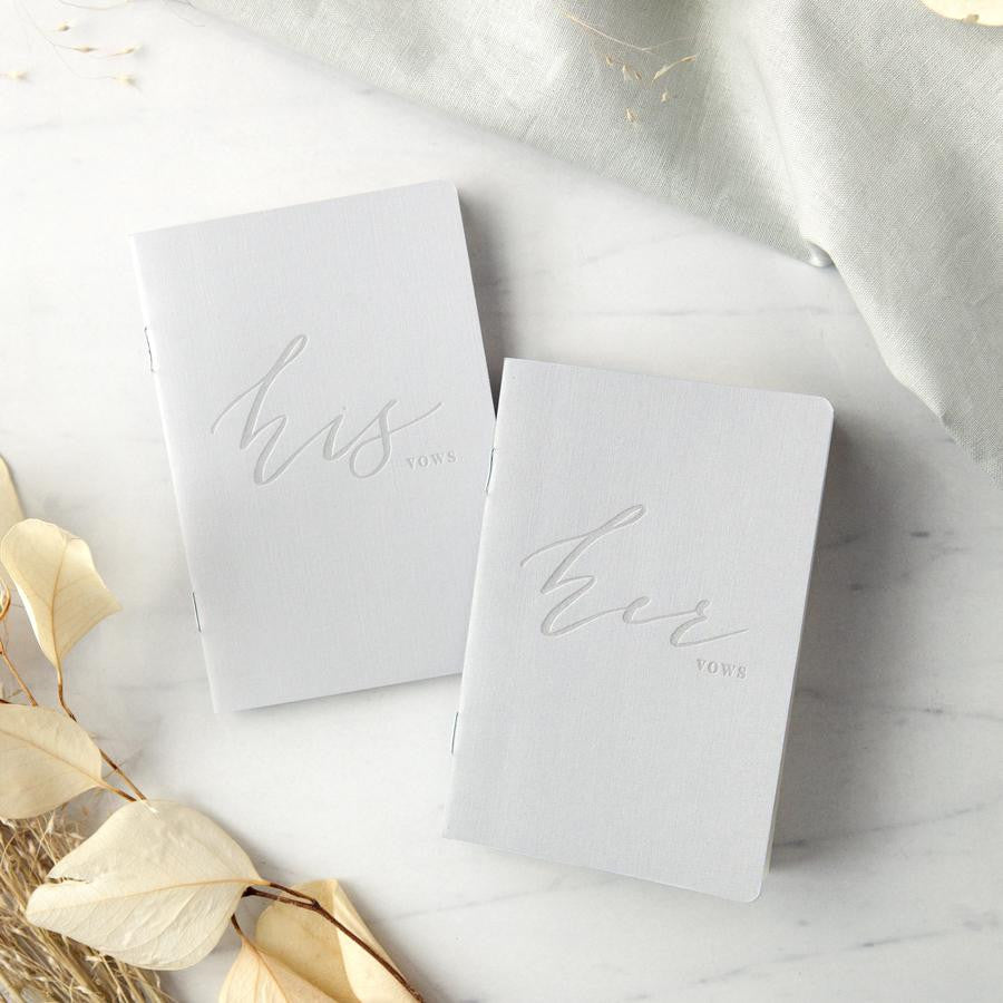 His and hers gray linen letterpressed vow books, featured in a marble-like background with flowers on one side and a linen cloth on the other.