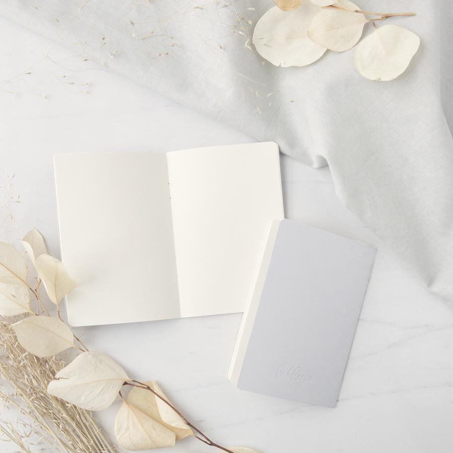 His and hers gray linen letterpressed vow books, one is opened and the other showing the back detail, featured in a marble-like background with flowers on one side and a linen cloth on the other.
