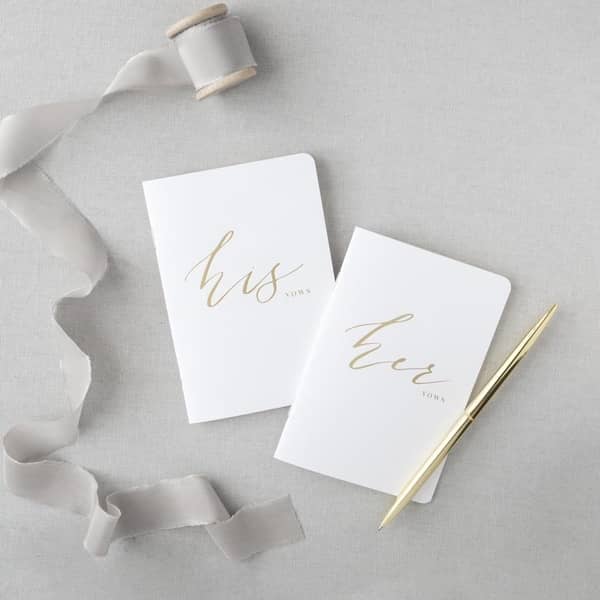 His and hers white, rounded-corner, gold-foil-stamped vow books featured in a gray background, with a gold pen on the right side.