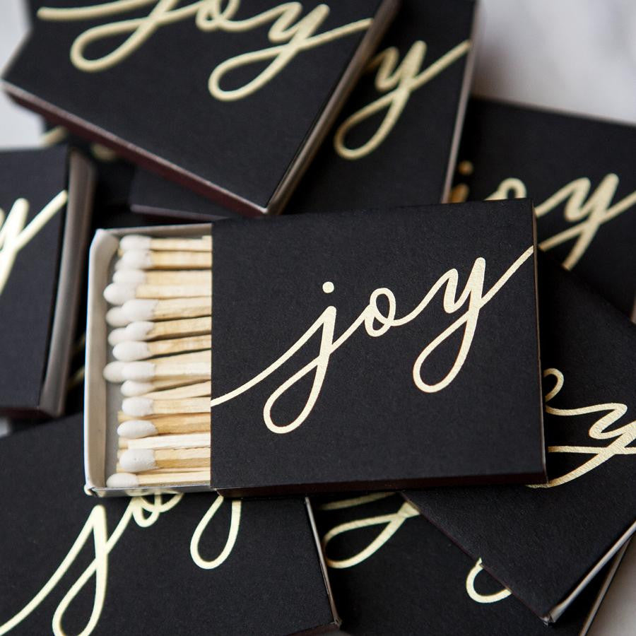 Joy metallic gold matchbox is featured,  black matchbox with the word Joy in gold metallic script lettering, the image displays several of them scattered on a white background, one in the center opened displaying the white tip matches inside. 