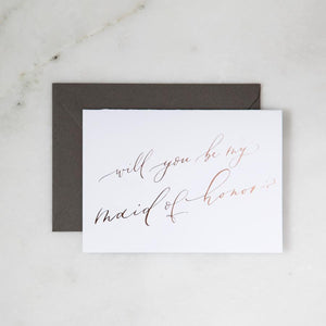 White envelope size card (approx. 3.5" x 5"), reads, "Will you be my maid of honor?" in rose gold text, with slate gray envelope option.