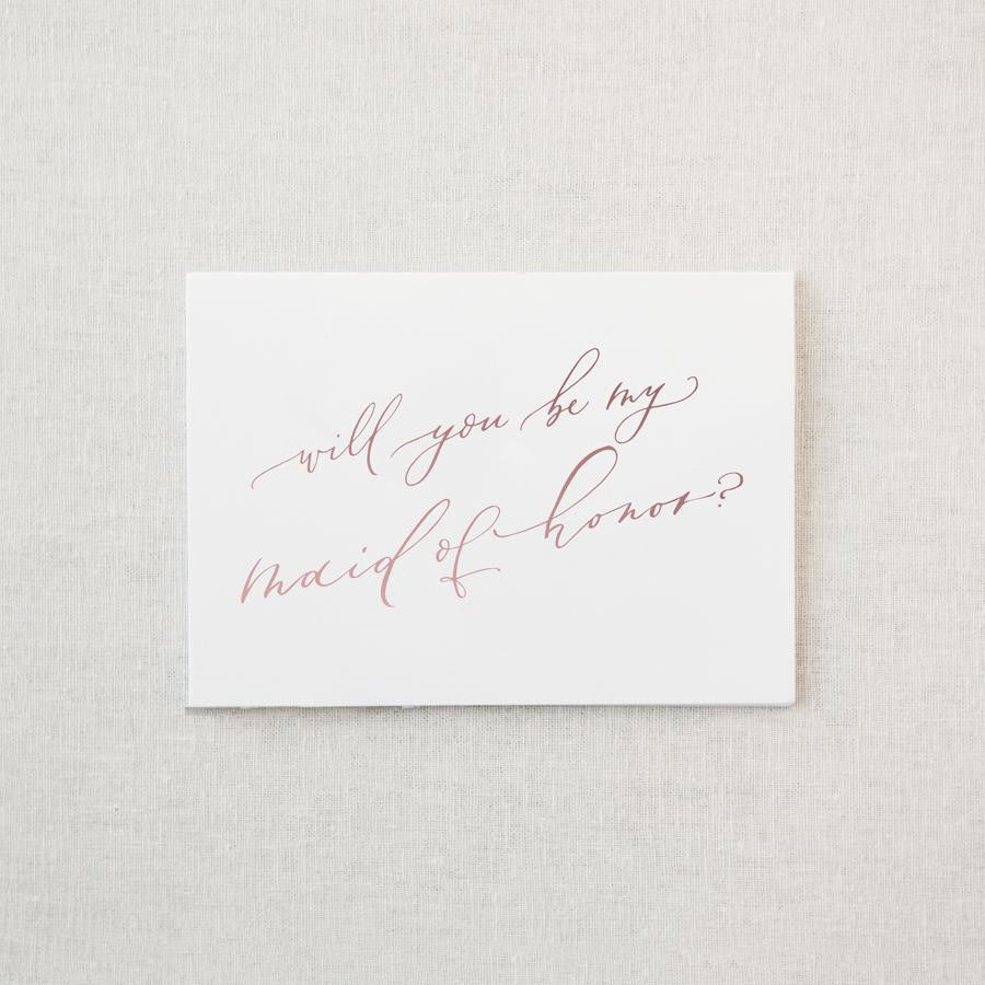 White envelope size card (approx. 3.5" x 5"), reads, "Will you be my maid of honor?" in rose gold text. 