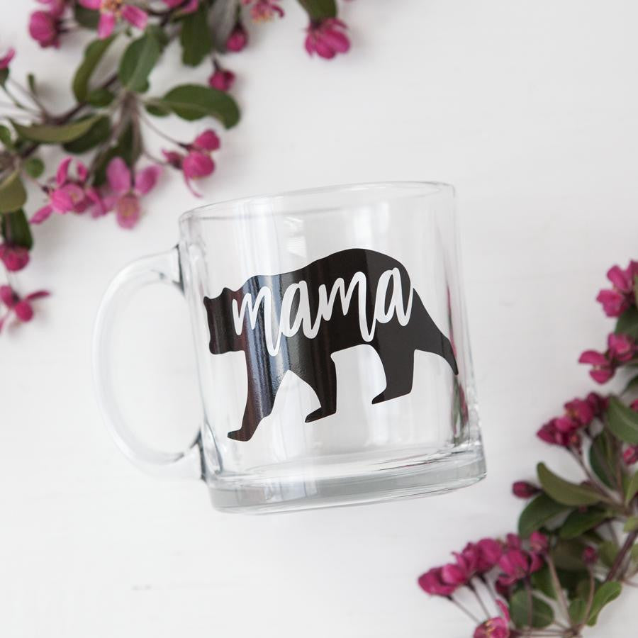 Mama bear mug featured: a transparent mug with a silhouette of a bear in black, cut out to reveal the mama bear in a white background with plants surrounding it. 