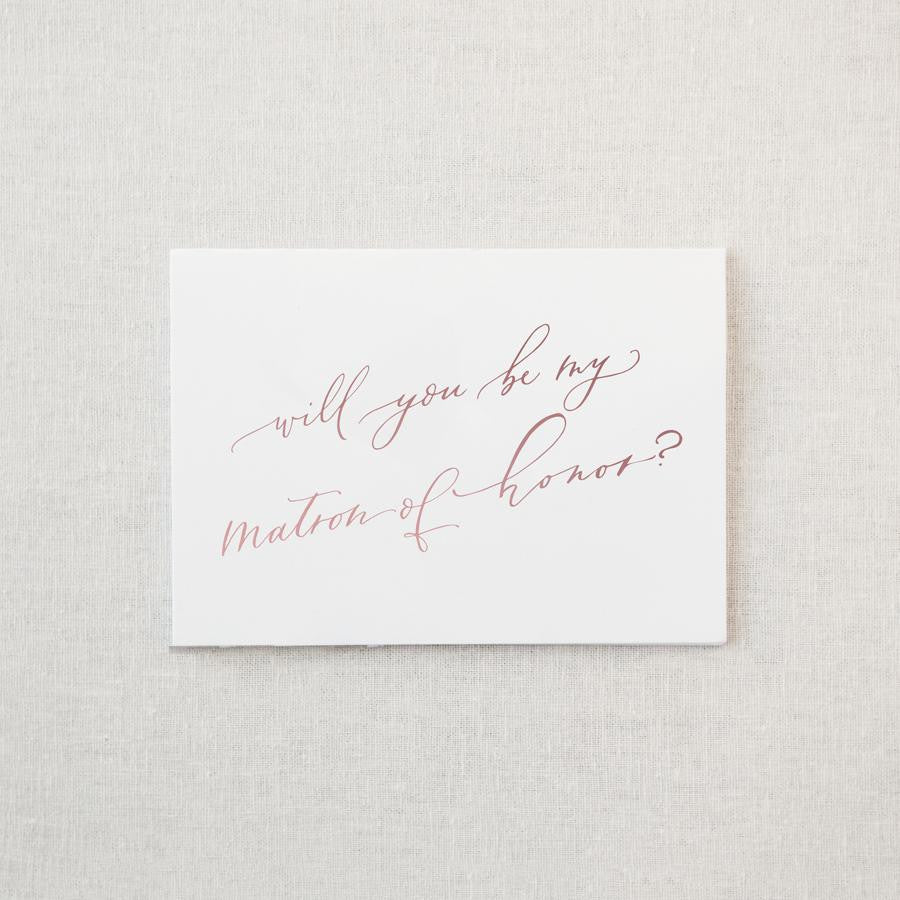 White envelope size card (approx. 3.5" x 5"), reads, "Will you be my matron of honor?" in rose gold text.