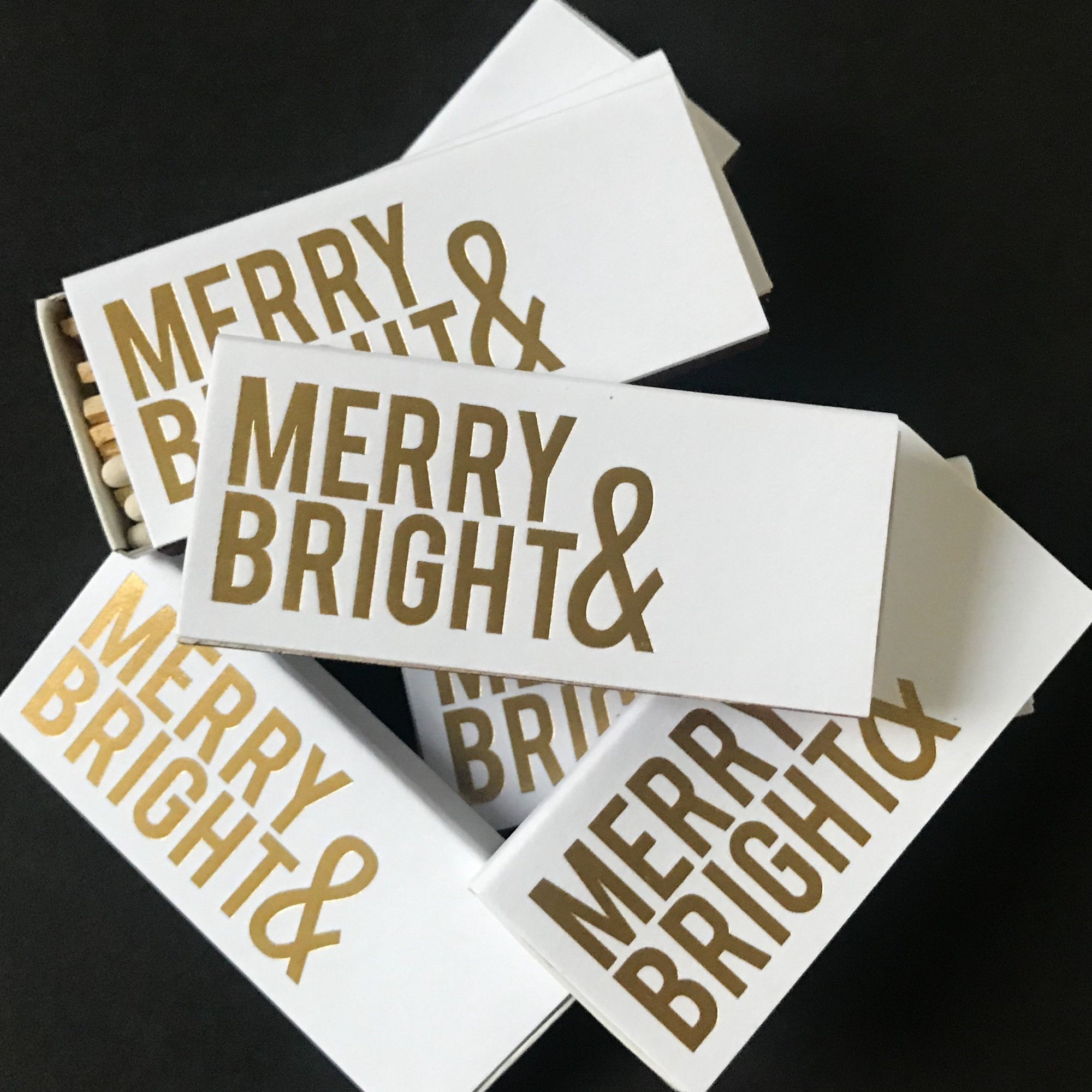 Merry and bright matchboxes, white rectangular matchboxes 6 of them with the letters Merry & Bright in gold foil. Ones is semi open showing the white tip matches inside of the matchbox. 