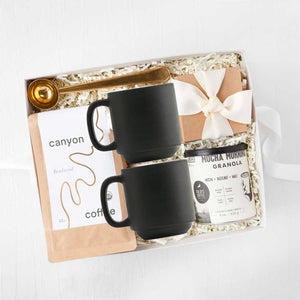 Gold clip coffee spoon, 'b toffee' with an ivory ribbon, Mocha morning granola in a cylinder can, 'canyon' coffee in a brown paper bag, and two black ceramic mugs, in ivory paper shread, in a gray 'foxblossm' box,