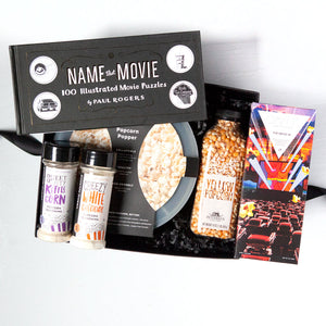 Black box with rectangular "name that movie - 100 illustrated movie puzzles by Paul rogers" book, two shakers of popcorn seasoning, one sweet and salty kettlecorn, the other cheezy white cheddar. Compartes :The Drive-in" chocolate bar, silicone popcorn popper bowl, bottle of popcorn kernels