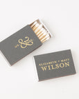 Mr & Mrs custom matchbox. The image showcases two personalized matchboxes, in chroma slate, one open and one closed. The open matchbox reveals a collection of white-tipped matches with “Mr & Mrs” printed in brushed gold foil on the cover. The closed matchbox displays the back side, featuring the names “Elizabeth and Matt Wilson” in brushed gold foil. Both are placed on a white background.