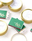 Mr and Mrs custom matchboxes shown in the image 4 of them chroma leaf personalized in shiny gold lettering with the letters Mr & Mrs compimented with personalized gold tin 4 oz candles that say Rachel & Ryan in different colors on a marble-like background. 