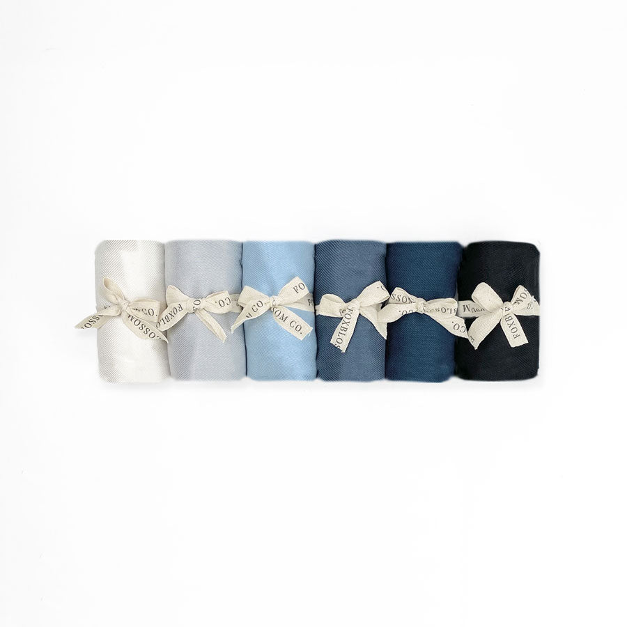 Pashmina Scarves, a row of rolled-up, tied-with-twine, various shades of pashmina scarves that come in the colors: light grey, ash grey, black, ivory, and navy.
