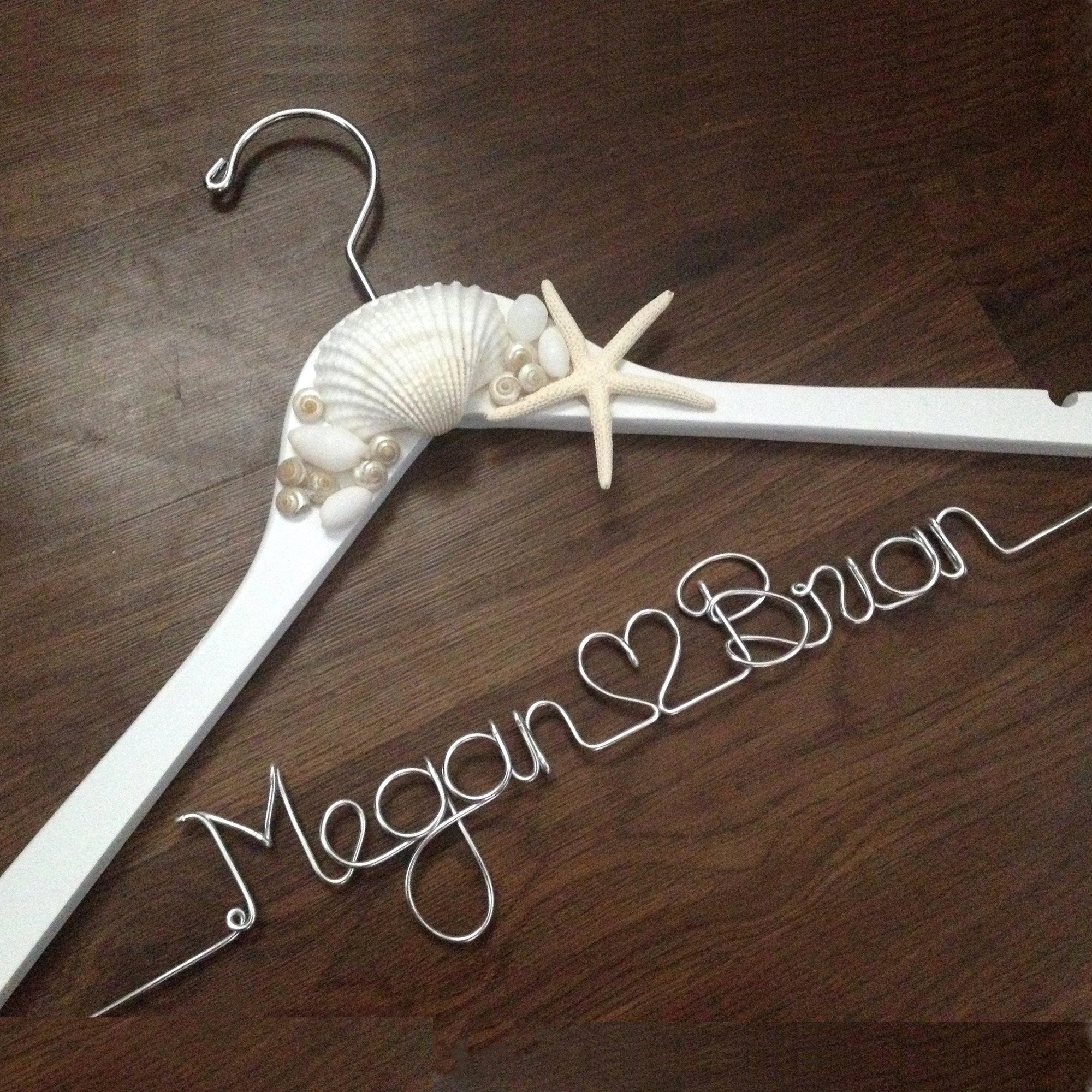 White hanger with shells and starfish accents. Personalized handcrafted wire hanger base in silver aluminum personalized with Megan, heart shape, and Brian on a dark wood background. 