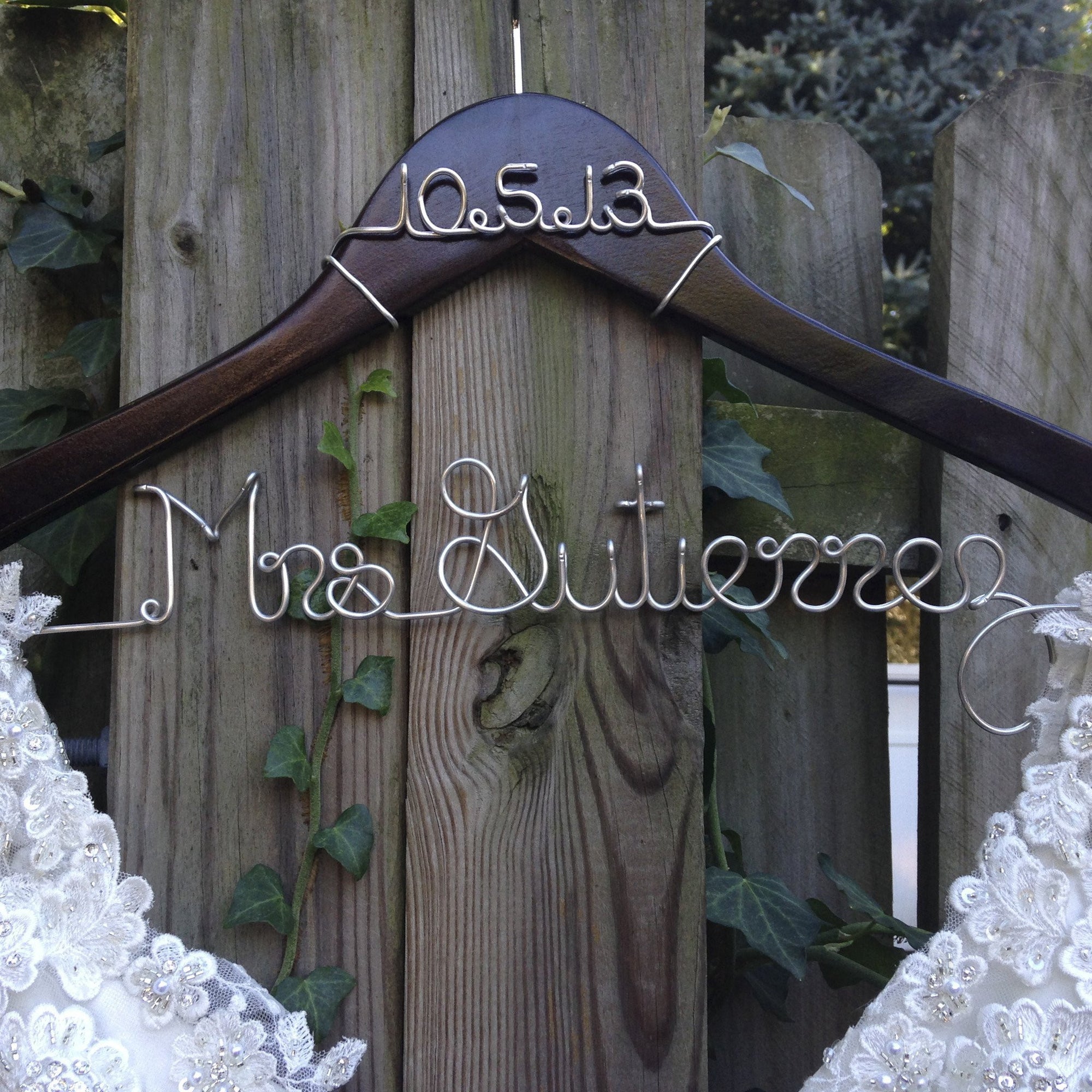 Dark wooden hanger with personalized date and base wire shaped into personalized name in silver aluminum wire with the date 10-5-13 and Mrs. Gutierrez on the bottom.   in front of a weathered wooden fence, partially obscured by green ivy. Holding a white wedding gown. 