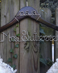 Dark wooden hanger with personalized date and base wire shaped into personalized name in silver aluminum wire with the date 10-5-13 and Mrs. Gutierrez on the bottom.   in front of a weathered wooden fence, partially obscured by green ivy. Holding a white wedding gown. 