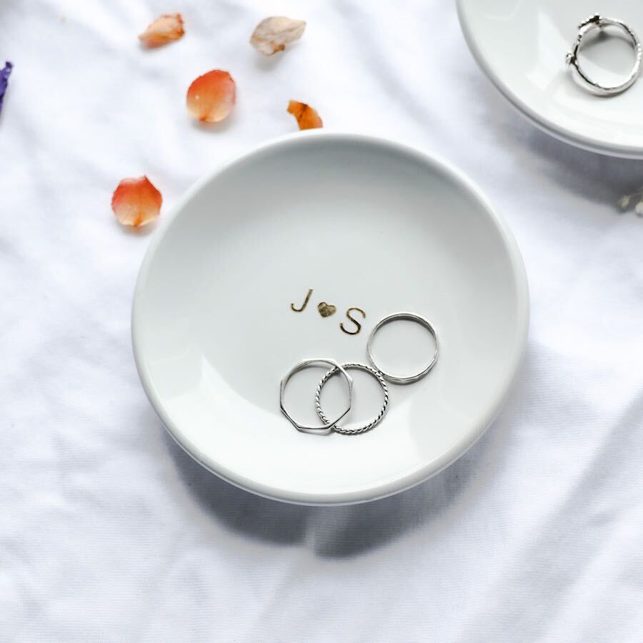 The image showcases our personalized  small, white ceramic ring dish with "J heart icon s" elegantly written in gold script, and  three silver ring resting inside.