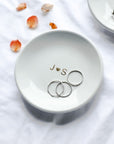 The image showcases our personalized  small, white ceramic ring dish with "J heart icon s" elegantly written in gold script, and  three silver ring resting inside.