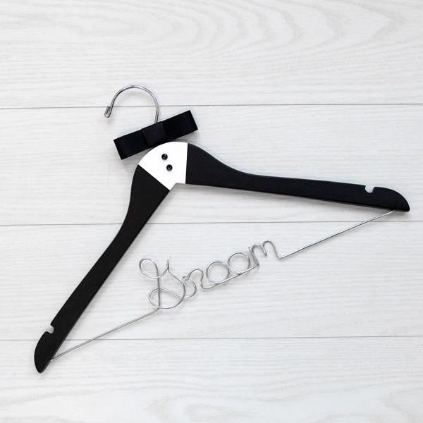 A luxe matte black hanger, hand-painted with a tuxedo design and finished with two tiny buttons and a grosgrain bowtie, customized with &quot;Groom,&quot; in silver wire with notches. On a white wooden background.