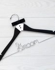 A luxe matte black hanger, hand-painted with a tuxedo design and finished with two tiny buttons and a grosgrain bowtie, customized with "Groom," in silver wire with notches. On a white wooden background.