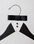 Close up of luxe matte black hanger, hand-painted with a tuxedo design and finished with two tiny buttons and a grosgrain bowtie with silver hock. Ona white background.